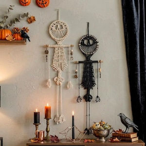 May include: Two macrame wall hangings, one in cream and one in black, featuring circular tops and elongated bodies suspended from wooden dowels. Each piece includes dangling cords with beads and charms, creating a symmetrical, intricate design.