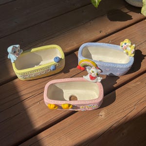 May include: Three rectangular ceramic planters shaped like bathtubs. One is yellow with a blue cat, one is light blue with a yellow cat, and one is pink with a white cat and a rainbow. Each planter features decorative embossed details and a drainage hole.