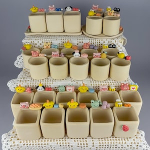 May include: A collection of small, cream-coloured ceramic cups arranged on tiered white lace doilies. Each cup features a unique, colourful miniature clay figurine attached to the rim, including animals like pigs, frogs, bees, and various food items.