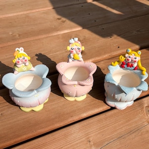 May include: Three ceramic flower-shaped planters, each featuring a small pig character on the rim. The left planter is blue and pink with a rabbit-eared pig, the centre is pink with a pig holding a banana, and the right is blue with a pig in a blonde wig.