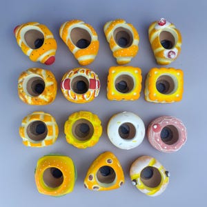 May include: A collection of fourteen ceramic food-shaped planters, each featuring a central circular opening with a dark mesh base. The assortment includes various yellow, orange, white, and pink designs resembling pastries, breads, and snacks.