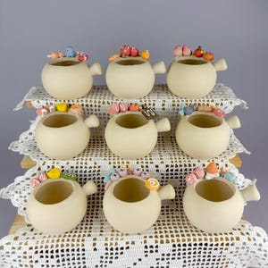 Peut inclure: Neuf tasses en céramique crème de forme ronde avec de petites anses, disposées sur trois rangées. Chaque tasse présente des décorations miniatures en argile colorée sur le bord, représentant diverses pâtisseries, fruits et sucreries.