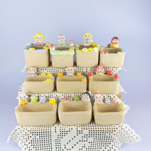 May include: A collection of ten small, square, beige planters arranged on a tiered display. Each planter features unique, colourful miniature animal figurines and food-themed decorations attached to the rims, including fruits, vegetables, and mushrooms.