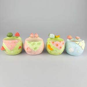 Peut inclure: Quatre petits pots de fleurs ronds en céramique avec des motifs de fruits. Chaque pot est peint dans des tons pastel avec un motif de bandana décoratif, des accents de fruits comme la pastèque, la pêche, le melon et le fruit du dragon.
