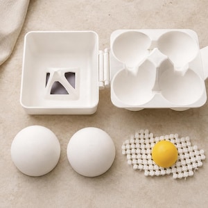 May include: A white plastic egg carton with four circular compartments and a hinged lid. Two smooth white spherical objects are placed below the carton, alongside a white crocheted piece featuring a single bright yellow sphere in the centre.