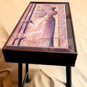 May include: A rectangular wooden side table with a dark brown finish and four slender legs. The tabletop features a glossy, inset decorative panel depicting a relief carving of an ancient Persian figure in profile, accented by a floral border.