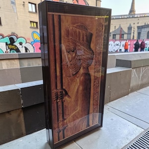 Può includere: Una teca rettangolare in legno marrone scuro con un pannello frontale in vetro. All'interno, una stampa in rilievo color seppia raffigura il profilo di un'antica figura con barba e un copricapo elaborato, che regge un bastone alto.