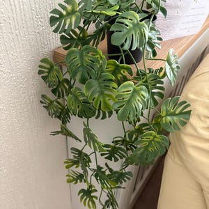 May include: A trailing artificial monstera plant with multiple green, split-leaf foliage stems cascading from a black plastic pot. The leaves feature characteristic perforations and notches, displaying a mix of deep and light green shades throughout.