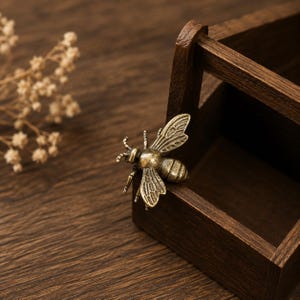 Peut inclure: Un objet d&eacute;coratif en m&eacute;tal en forme d'abeille avec une finition en laiton antique, pr&eacute;sentant des ailes d&eacute;taill&eacute;es et un corps segment&eacute;, pos&eacute; sur le bord d'une petite bo&icirc;te en bois ouverte avec une poign&eacute;e.