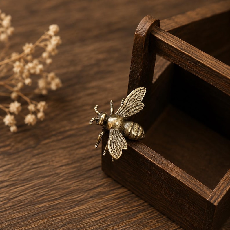 Peut inclure: Un objet d&eacute;coratif en m&eacute;tal en forme d'abeille avec une finition en laiton antique, pr&eacute;sentant des ailes d&eacute;taill&eacute;es et un corps segment&eacute;, pos&eacute; sur le bord d'une petite bo&icirc;te en bois ouverte avec une poign&eacute;e.
