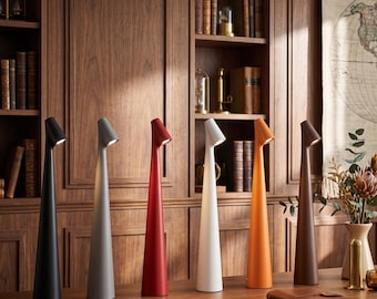 Peut inclure: Six lampes de table hautes et fines sont alignées sur une surface en bois. Les lampes présentent une base conique et une petite tête lumineuse inclinée au sommet. Les couleurs de gauche à droite sont noir mat, gris, rouge, blanc, orange et brun.
