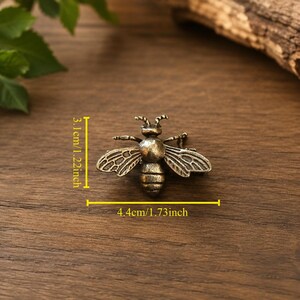 Peut inclure: Un ornement en m&eacute;tal en forme d'abeille de couleur bronze avec des motifs d'ailes d&eacute;taill&eacute;s et un corps segment&eacute;. Des lignes de mesure jaunes indiquent une hauteur de 3,1 cm et une largeur de 4,4 cm, offrant une &eacute;chelle claire pour cet objet.