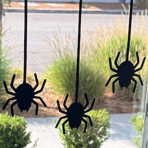 May include: Five black spider silhouettes hanging from vertical black lines. Each spider features a rounded body and eight distinct legs. The spiders are arranged in a row, suspended against a clear surface, creating a uniform decorative display.