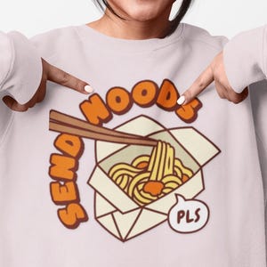 May include: A light pink crewneck sweatshirt featuring a graphic illustration of a white takeout container filled with yellow noodles and brown chopsticks. The text "SEND NOODS" is printed in orange above the container, with "PLS" in a bubble.