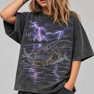 Może przedstawiać: Ciemnoszary t-shirt z krótkim rękawem z nadrukiem graficznym przedstawiającym aligatora wyłaniającego się z ciemnej wody. Scena obejmuje bagnisty krajobraz z cyprysami i jasnofioletowymi błyskawicami na zachmurzonym niebie.