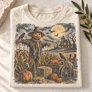 May include: A cream-colored t-shirt featuring a graphic print of a scarecrow with a pumpkin head standing in a cornfield. The scene includes a wooden cart with pumpkins, a haunted house, a full moon, and black crows under a dark, cloudy sky.