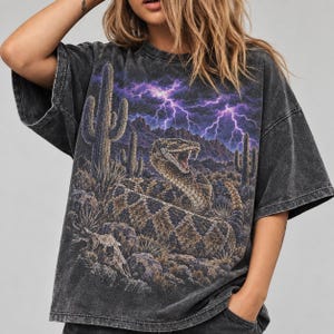 Può includere: Una t-shirt grigio antracite con effetto lavaggio acido, caratterizzata da una stampa grafica di un serpente a sonagli avvolto con la bocca aperta, in un paesaggio desertico con cactus, rocce e un teschio sotto un cielo viola con fulmini.