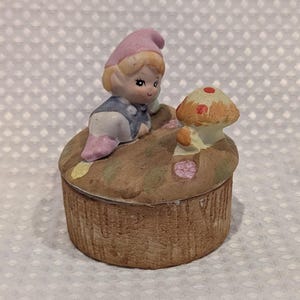 May include: A ceramic trinket box featuring a sculpted gnome wearing a pink hat and blue outfit, sitting on a brown textured base next to a small tan mushroom with red spots. The lid is round and painted with earthy tones and small floral details.