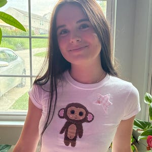 May include: A white short-sleeved t-shirt featuring a brown monkey applique with pink accents on the ears and cheeks, positioned next to a light pink and white gingham star applique on the upper right side of the chest area.
