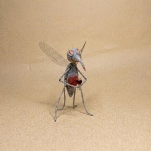 Op de afbeelding: Een naaldgevilte muggenfiguur met een grijs lichaam, een opvallend rood achterlijf en grote ogen. Het insect heeft dunne, draadachtige poten en doorschijnende vleugels en houdt een klein, helder glas vast met een donkerrode vloeistof.