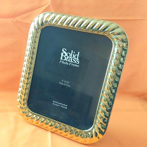 May include: A gold-colored solid brass photo frame with a twisted rope-style border. The black insert features the text Solid Brass Photo Frame, 8 inches by 10 inches, and hand-polished lacquer finish. The frame has a square shape with rounded corners.