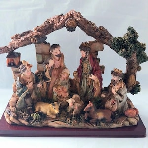 May include: A nativity scene figurine set featuring a brown textured stable structure with a tree. The scene includes figures of adults and a child in robes, a sheep, and a small horse, all rendered in muted earth tones on a dark red rectangular base.
