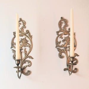 May include: A pair of wall-mounted candle sconces made of polished brass with intricate scrollwork and leaf-like patterns. Each sconce features a single metal cup holding a long, slender, off-white taper candle, creating a symmetrical display.