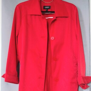 May include: A bright red long-sleeve coat featuring a pointed collar, a front button closure with three visible buttons, and cuffed sleeves. The interior neckline displays a black label with the text COMPANY and a gold-toned chain hanging loop.