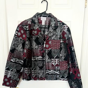 May include: A long-sleeve zip-up jacket featuring a patchwork pattern in black, deep red, and silver. The design includes various geometric and ornate motifs across the fabric. It has a standard collar, a silver-toned zipper, and long sleeves.