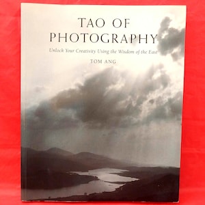 May include: A softcover book titled Tao of Photography: Unlock Your Creativity Using the Wisdom of the East by Tom Ang. The cover features a black and white landscape photograph of a lake surrounded by hills under a cloudy, dramatic sky.