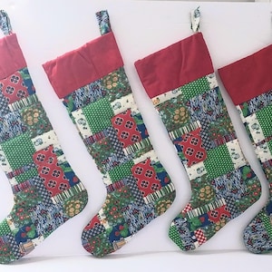 Puede incluir: Cuatro calcetines navideños con un diseño de patchwork de varios cuadrados de tela en patrones rojos, verdes, azules y blancos. Cada calcetín tiene un puño rojo sólido en la parte superior y un lazo de tela para colgar, dispuestos en fila.
