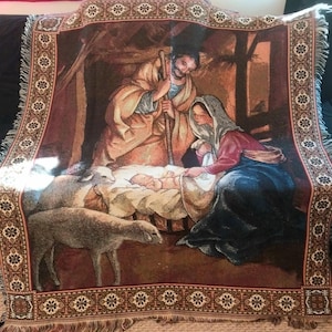 May include: A woven tapestry throw blanket featuring a nativity scene with a central figure in tan robes, a figure in a red and blue garment with a head covering, and an infant in a manger. Two sheep are visible in the foreground with fringe edges.