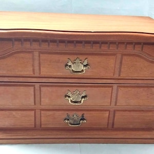 May include: A wooden jewelry box with a rectangular shape, featuring a top lid and two lower drawers. The exterior is a medium brown wood grain finish with decorative molding and three brass-colored metal drawer pulls centered on the front.