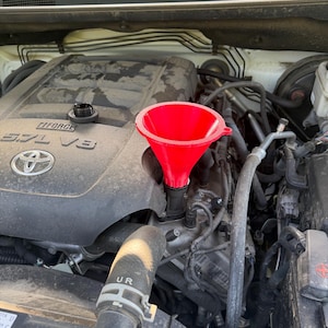 May include: A bright red plastic funnel inserted into the oil fill port of a dusty Toyota 5.7L V8 engine. The black oil cap sits nearby on the engine cover, which features the Toyota logo and the text i-FORCE 5.7L V8 in raised silver lettering.