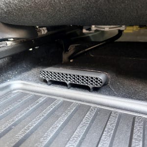 May include: A black plastic air vent cover with a honeycomb mesh pattern installed on the floor of a vehicle, positioned directly in front of a black rubber floor mat beneath the seat assembly and various internal car components.