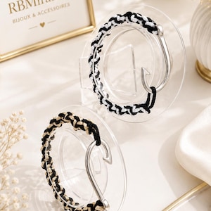 Peut inclure: Deux bracelets dotés d'un fermoir en métal argenté en forme d'hameçon. L'un des bracelets présente un cordon tressé noir et blanc, tandis que l'autre est composé d'un cordon tressé noir et doré. Chaque bracelet est sur un support circulaire.