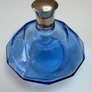 May include: A cobalt blue glass perfume bottle with a faceted, multi-sided body and a smooth, rounded metallic silver-toned stopper. The vessel features a wide, low-profile base that tapers upward toward the narrow neck and the reflective cap.