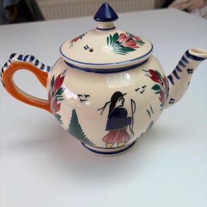 May include: A cream-colored ceramic teapot featuring hand-painted illustrations of a person with a bow, green trees, and red floral motifs. The teapot has a bright orange handle with blue stripes, a blue-trimmed lid, and a matching blue-striped spout.