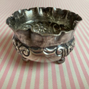 May include: A small, antique silver-colored bowl with a ruffled rim and ornate floral and scrollwork detailing. The bowl rests on three small feet and is likely used for serving small portions or as a decorative piece.