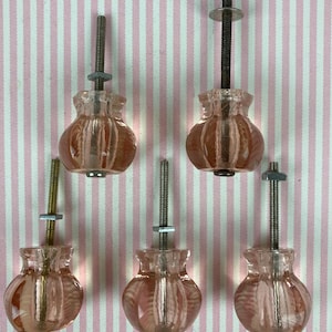 May include: Five translucent pink glass cabinet knobs with a rounded, fluted shape. Each knob features a central metal bolt extending from the top, equipped with a small metal nut and washer for secure installation on furniture or cabinetry.
