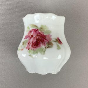 May include: A small, white porcelain vase with a scalloped rim. The vase features a hand-painted design of a large, pink rose with green leaves. The vase has a glossy finish and a slightly curved shape.