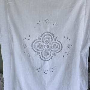 May include: White tablecloth with a central floral cut-out design. The cut-out is surrounded by smaller floral and leaf-shaped cut-outs. The edges of the tablecloth have a scalloped trim.