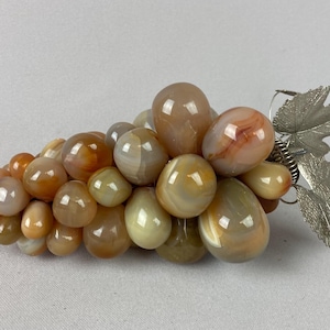 May include: A decorative cluster of faux grapes made from polished stones in shades of brown, orange, and cream. The grapes are attached to a silver-colored metal stem with leaves, creating a unique home decor item.