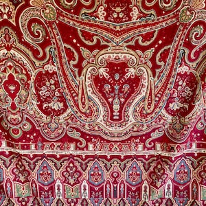 May include: A close-up of a red textile with an ornate paisley pattern. The design features swirling floral and geometric elements in shades of gold, green, and blue. The fabric has a velvety texture and a decorative border.