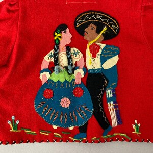 以下が含まれることがあります： 赤いフェルトジャケットに、伝統的なメキシコ衣装を着た男性と女性の刺繍が施されています。女性は花柄の刺繍が施された青いスカートを、男性は黒の帽子をかぶった青と白のシャツを着ています。