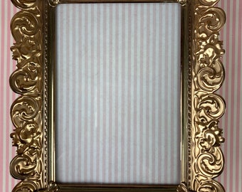May include: A gold-colored ornate picture frame with a detailed, intricate design. The frame is made of metal and has a rectangular shape.