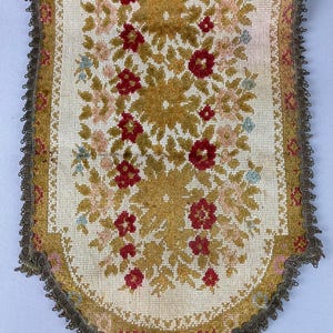 May include: A rectangular textile runner with a rounded bottom edge, featuring a needlepoint floral pattern in shades of mustard yellow, deep red, and light pink on a cream background, finished with a dark brown decorative braided trim.