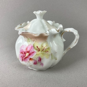 May include: A white porcelain sugar bowl with a lid, featuring a floral design in pink and yellow. The bowl has a handle and is trimmed with gold accents. The lid has a decorative finial.