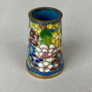 May include: A cylindrical thimble with a floral design. The thimble features a brass rim and base, with a blue interior. The exterior is decorated with colorful flowers and leaves on a light green background.