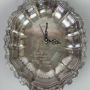May include: A silver-colored decorative clock with ornate detailing around the rim. The clock face is a shallow dish with the words "HOW DID IT GET SO LATE SO SOON?" The clock hands are black.